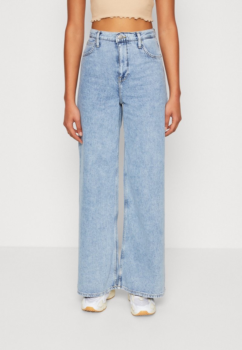Personne portant un jean bleu clair taille haute à jambes larges, un crop top beige et des baskets blanches, debout sur un fond blanc.