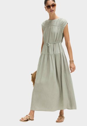 Femme portant une robe maxi sans manches vert clair avec taille à cordon, lunettes de soleil écaille, sandales dorées et tenant un sac à franges marron.