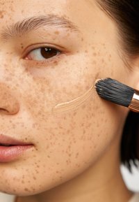 Un gros plan sur un visage présentant une fondation marron clair appliquée au pinceau sur une peau avec des taches de rousseur prononcées et une texture lisse.