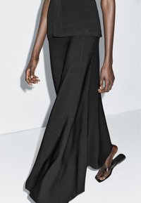 Person trägt schwarze Hose mit weitem Bein und schwarze Flip-Flop-Sandalen, geht vor einem schlichten weißen Hintergrund.