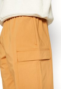 Pantalon cargo marron clair avec poche à rabat latérale et taille élastique, associé à l'ourlet d'un sweat beige visible au-dessus.