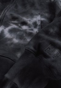 Sweatshirt preta com padrão tie-dye em cinza, textura macia de algodão, fecho de correr e logótipo "GAP" em relevo no punho.