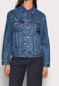 Keskisininen farkkutakki klassisella kauluksella, napillinen etumus, kaksi rintataskua ja sivutaskut. Korostaa vastaväriä ompeleissa.