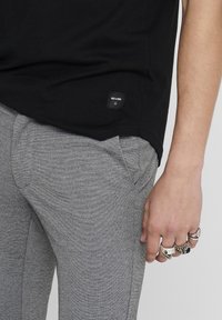 Schwarzes Baumwoll-T-Shirt mit kleinem Etikett; graue strukturierte Hose mit Seitentaschen. Hand geschmückt mit mehreren Silberringen.