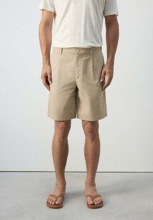 Hombre con shorts beige, camiseta blanca y sandalias marrones, de pie sobre un suelo gris frente a una pared blanca lisa.