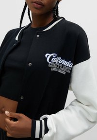 Black and white varsity jacket with a snap button closure and ribbed cuffs, featuring "California Coastline Laguna Beach" embroidery.