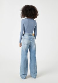 Langärmliges, geripptes, hellblaues Oberteil, kombiniert mit hochgeschnittenen, weiten, blauen Jeans mit einem braunen Lederpatch am Rücken.