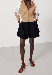 Beżowy zamek-hiphoker z białymi rękawami, zestawiony z czarnymi, luźnymi szortami. Całość uzupełniają czerwone sandały na paski. Gładkie tekstury.