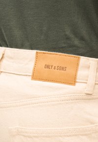 Tan lædermærke med "ONLY & SONS"-branding på cremefarvede jeans, båret med mørkegrøn top.