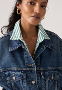 Γυναίκα με χρυσούς κρίκους στα αυτιά, που φοράει πράσινο και λευκό ριγέ πουκάμισο με γιακά κάτω από μπλε τζιν μπουφάν.