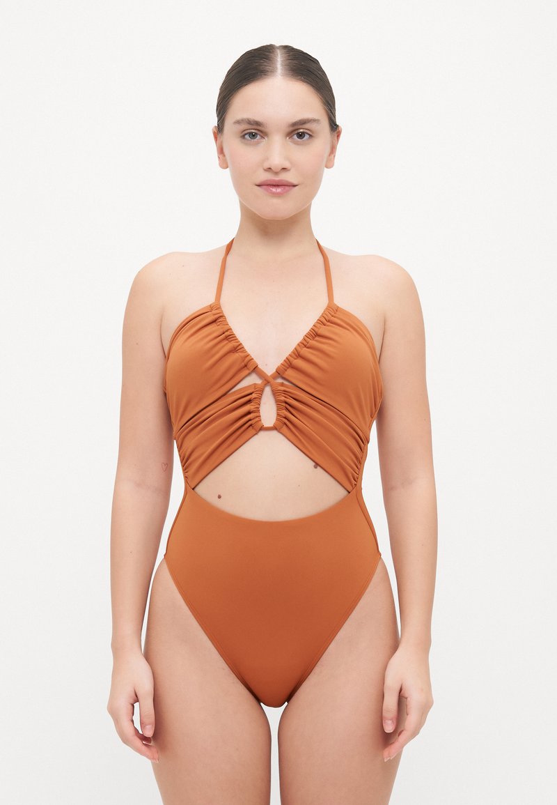 Maillot de bain une pièce orange brûlé avec un design froncé, des bretelles à nouer autour du cou et une découpe ovale à la taille. Texture de tissu lisse.
