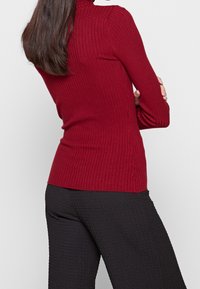 Pull à col roulé côtelé de couleur rouge profond, coupe ajustée, texture douce. Assorti à un pantalon noir texturé. Mise en valeur des détails du vêtement.