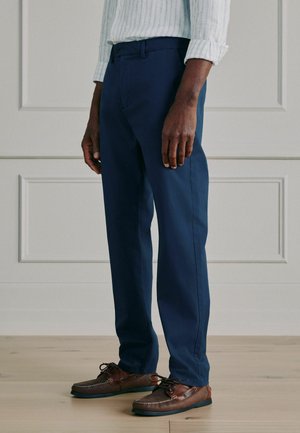 Man wearing navy blue trousers, brown leather boat shoes, and a white long-sleeve shirt with thin blue stripes, standing indoors.