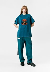 Camiseta de manga corta en color azul verdoso con un diseño bordado de oso de peluche marrón. Combinada con pantalones azules verdosos a juego y calzado negro texturizado.