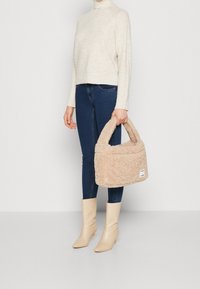 Person in beigem Rollkragenpullover, dunkelblauen Skinny-Jeans, beigen Absatzstiefeln, die eine beigefarbene, flauschige Handtasche an der Seite hält.