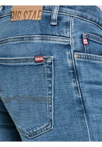 Mėlyno džinsinio audinio kelnų užpakalinės kišenės artimas planas su rudomis Big Star odinėmis etikete ir raudonomis audinių etiketėmis ant kišenių.