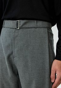 Grijze broek van gestructureerde stof, met een plat voorpand, riemlussen en een metalen gesp. Close-upweergave benadrukt details en stiksels.