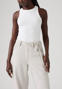 Bijela rebrasta majica bez rukava s okruglim izrezom, uparena s svijetlo bež hlačama visokog struka koje imaju pojas za vezanje i prednje džepove.