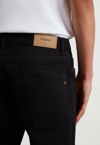 Pantaloni in denim neri con etichetta in pelle contrassegnata "FARAH", con tasche posteriori standard e dettagli dorati discreti. Tessuto liscio.