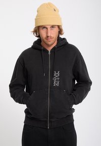 Homme portant un bonnet beige en maille et un sweat à capuche noir zippé avec un texte blanc vertical sur la poitrine, mains dans les poches avant, regardant devant.