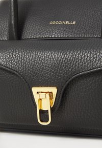 Black leather handbag with a textured finish, gold hardware, and a distinctive latch closure, featuring embossed branding on the front.