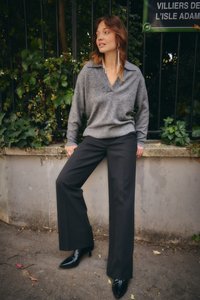 Ženska, oblečena v sivi pulover, črne široke hlače in črne škornje z visokimi petami, se naslanja na nizko kamnito steno, za njo pa je zelena rastlinje.