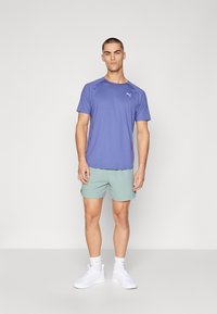 Herre atletisk tøj bestående af en lilla kortærmet skjorte og lysegrønne shorts, parret med hvide sneakers. Simpelt og moderne design.