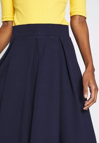 Laivastonsininen laskostettu hame, joka on valmistettu joustavasta kankaasta, leveällä vyötärönauhalla, sileällä pinnalla ja hulmuavalla siluetilla.