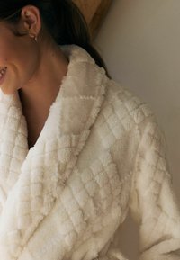 Krémový koupací plášť vyrobený z měkké, strukturované tkaniny, s výrazným vzorem v podobě diamantů a šálovým límcem pro větší teplo.