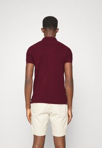 Man med kort hår som vänder bort sig, iklädd en vinröd polo och beige shorts mot en ren vit bakgrund.