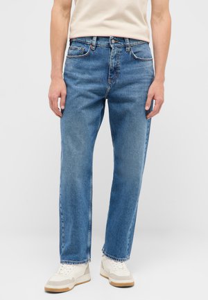 Person wearing blue straight-leg jeans, white sneakers, and a tucked-in beige t-shirt against a plain background.