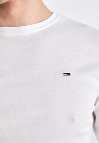 Biała bawełniana koszula z długim rękawem, z okrągłym dekoltem i małym haftowanym logo z czerwonymi, białymi i niebieskimi paskami na piersi.
