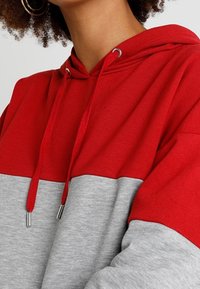 Felpa con cappuccio a blocchi di colore rosso e grigio, con cappuccio regolabile da coulisse, dettagli in metallo e tessuto morbido e testurizzato.