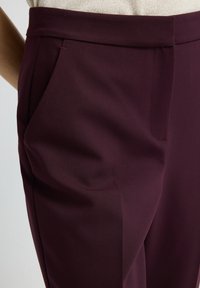 Close-up van donkerpaarse op maat gemaakte broek met zijzakken, gedragen met een beige top op een persoon.
