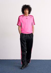 Rozā Adidas sporta krekls ar melniem un rozā akcentu svītrām, kombinēts ar melnām sporta biksēm ar sānu svītrām un tumšām kurpēm.