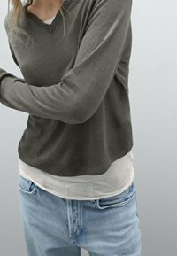 Maglione grigio a maglia con scollo a V, indossato sopra una maglietta bianca. Abbinato a jeans azzurri chiari. Tessuto morbido e vestibilità comoda.