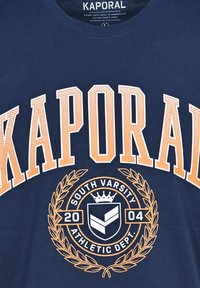 Camiseta de algodón azul marino con un texto en negrita naranja "KAPORAL" y un gráfico circular que incluye "SOUTH VARSITY" y un diseño de laurel.