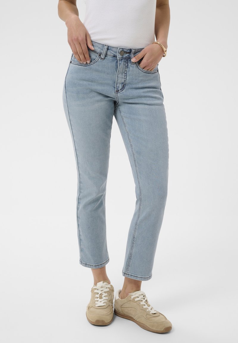 Femme portant un jean court bleu clair, un haut blanc sans manches, des baskets beiges et un bracelet en or, mains dans les poches avant.