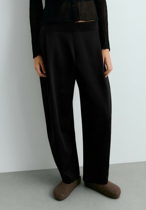 Personne portant un pantalon noir ample, un haut noir texturé à manches longues et des chaussons marron à enfiler, debout à l'intérieur.