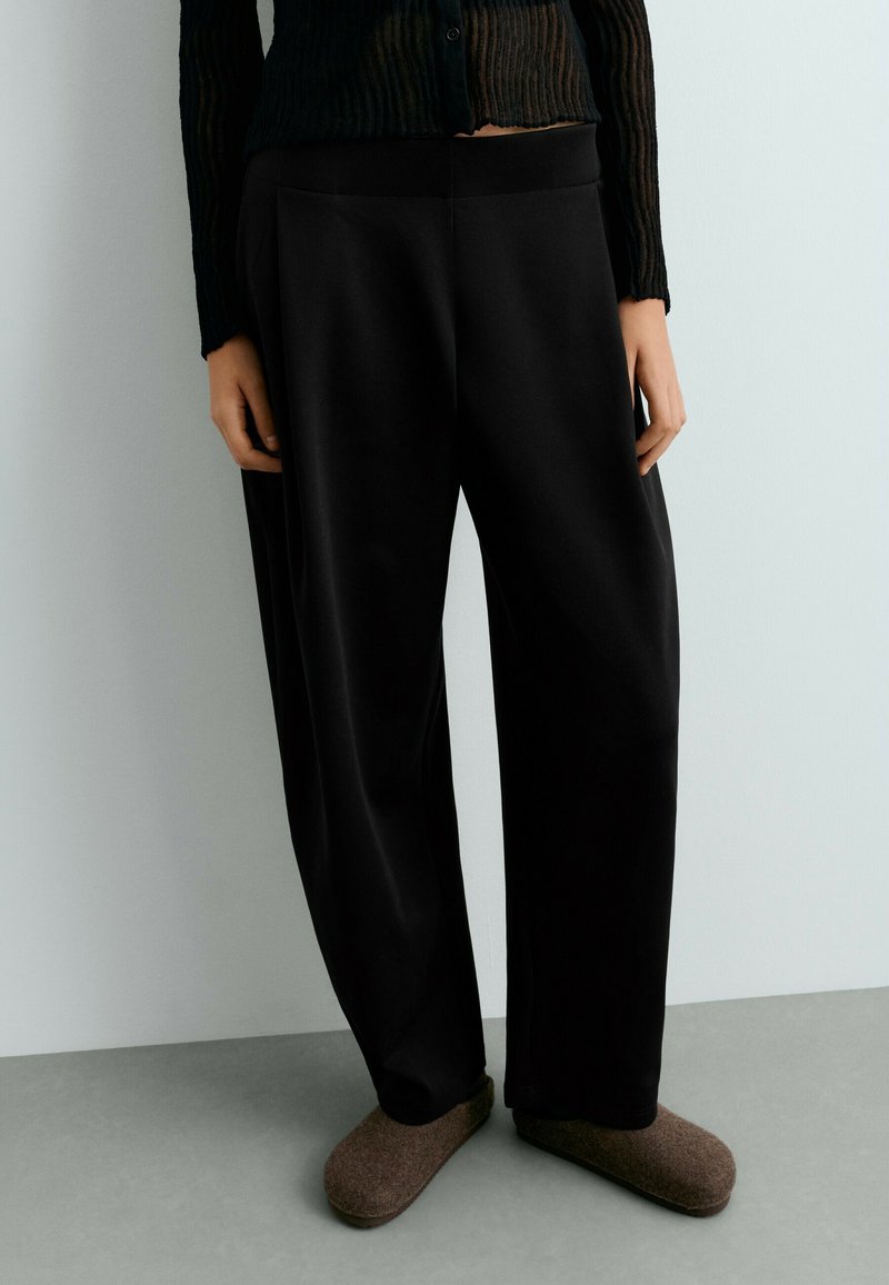 Personne portant un pantalon noir ample, un haut noir texturé à manches longues et des chaussons marron à enfiler, debout à l'intérieur.