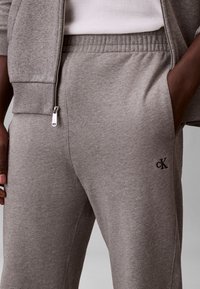 Close-up of person wearing light grey sweatpants with Calvin Klein logo, paired with matching zip-up hoodie and white ribbed shirt.