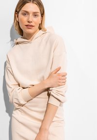 Hellbeige Kapuzenpullover aus weichem Stoff mit einer großen Kapuze und gerippten Bündchen, getragen über einem passenden, figurnahen Rock. Einfaches Design, minimale Details.