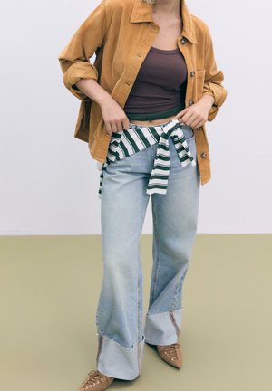 Persona con una chaqueta de pana mostaza, camiseta sin mangas marrón, pantalones vaqueros anchos de pierna clara, camisa de rayas anudada a la cintura y zapatos marrones puntiagudos.