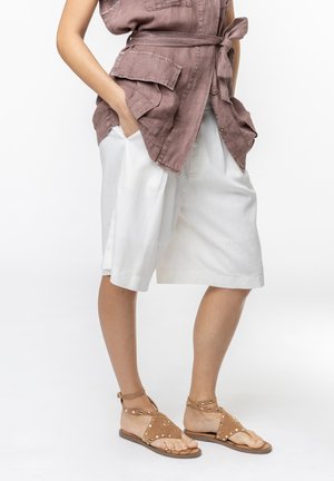 Donna con pantaloncini bianchi fino al ginocchio, gilet marrone con cintura e grandi tasche, e sandali bassi marroni con borchie.