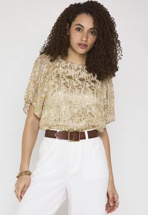 Femme aux cheveux bouclés portant un chemisier beige à motifs, un pantalon blanc taille haute avec une ceinture marron, et des bracelets dorés sur fond blanc.