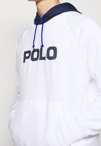 Sudadera blanca con capucha azul marino, detalles en azul y un gran logo "POLO" en negro en el frente. Con bolsillo canguro y puños de canalé.