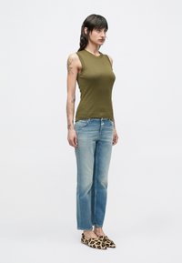 Mujer de cabello negro que lleva una blusa sin mangas color verde oliva, jeans azul claro y zapatos planos con estampado de leopardo, de pie frente a un fondo liso.