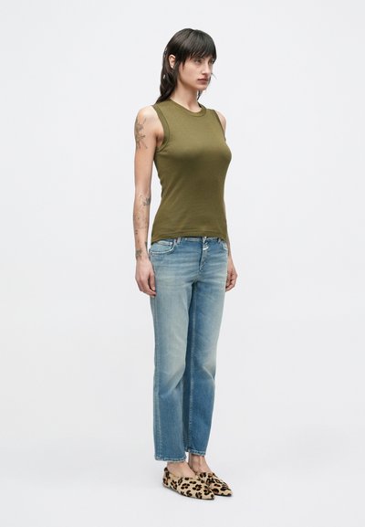 Γυναίκα με μαύρα μαλλιά που φοράει αμάνικο τοπ σε ελιά αποχρώσεις, ανοιχτόχρωμα μπλε τζιν και φλατ παπούτσια με λεοπάρ σχέδιο, στέκεται μπροστά σε απλό φόντο.