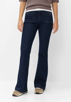 Kvinde iført mørkeblå bootcut-jeans, beige sneakers og en brun sweater, stående mod en hvid baggrund.