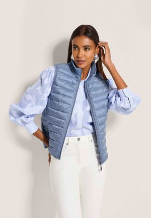 Frau mit langen dunklen Haaren trägt eine hellblaue, geblümte Bluse mit Puffärmeln, eine hellblaue gesteppte Weste, weiße Hose und silberne Ohrringe und posiert seitlich.